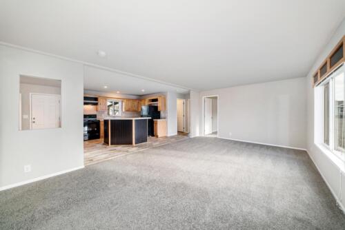 An empty living room with gray carpet and a kitchen.